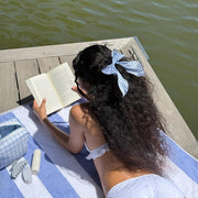 model reading a book while wearing Bow Barrette in Baby Blue Gingham in hair and laying on striped towel across wooden dock next to emi jay hair care and accessories 